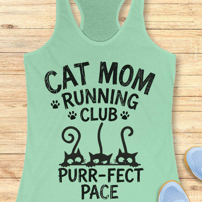 A mint green racerback tank top with a black graphic print that reads 'CAT MOM RUNNING CLUB PURR-FACT PACE' with cat silhouettes and paw prints, laid flat on a light wood background with partial shoes visible at the edges.