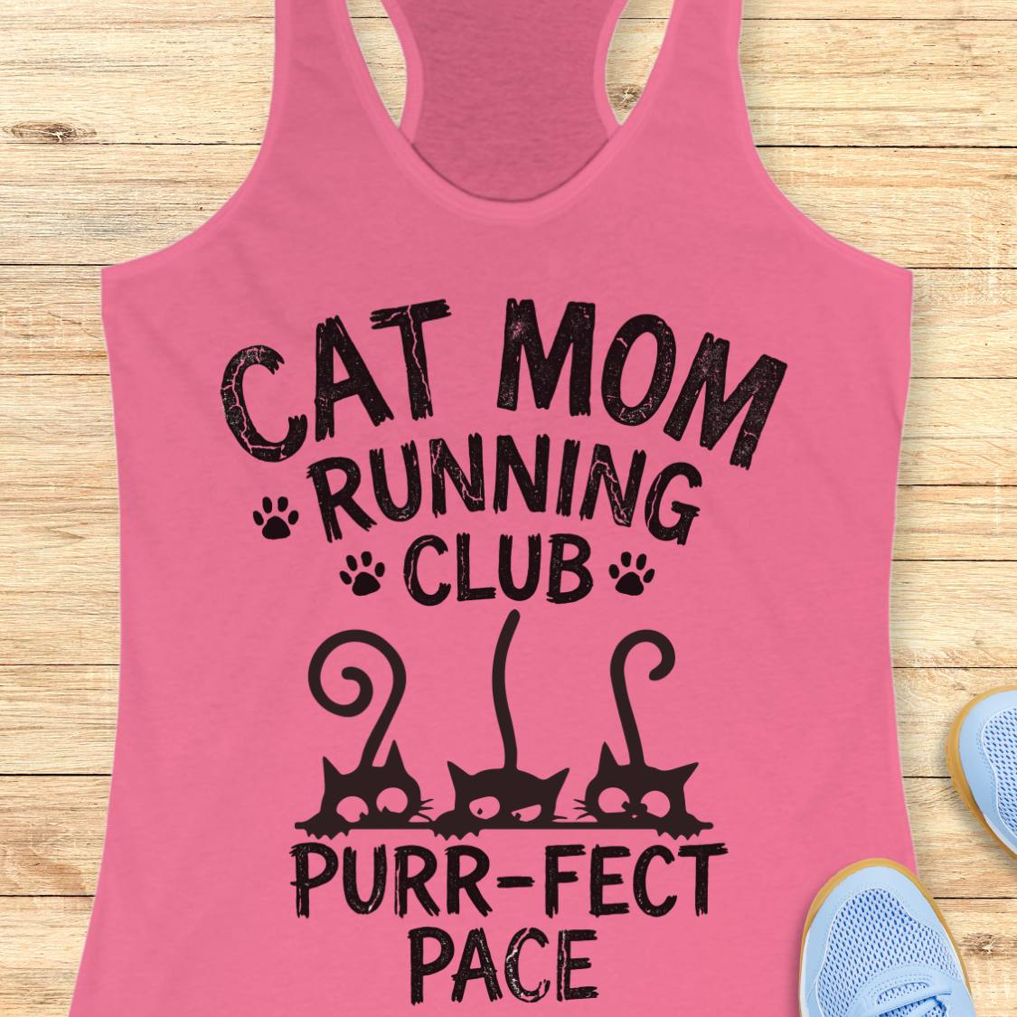 A pink racerback tank top laid flat on a light wood surface, printed with the phrase 'CAT MOM RUNNING CLUB PURR-FACT PACE' in black text with cat silhouette graphics. Partial blue sneakers are visible at the edges as styling props.