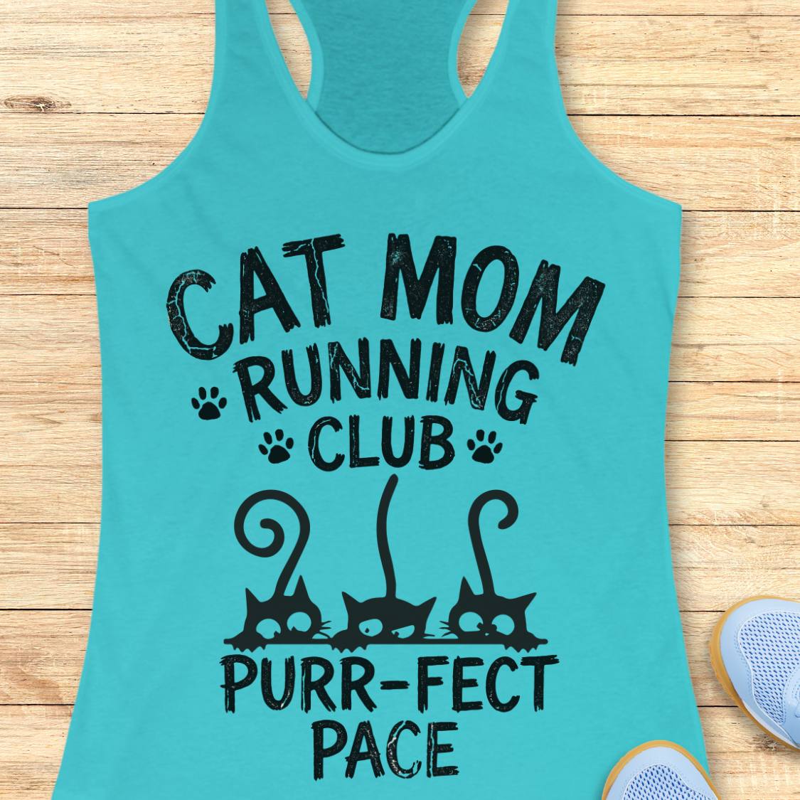 A turquoise racerback tank top laid flat on a light wood surface, printed with the phrase 'CAT MOM RUNNING CLUB PURR-FECT PACE' in black lettering with paw prints and cat silhouettes. Part of a light blue shoe is visible at the bottom right.