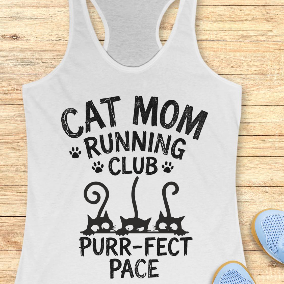 A white racerback tank top laid flat on a light wood surface, printed with the phrase 'CAT MOM RUNNING CLUB PURR-FECT PACE' and three black cat silhouettes. Part of a blue shoe is visible at the lower right edge.