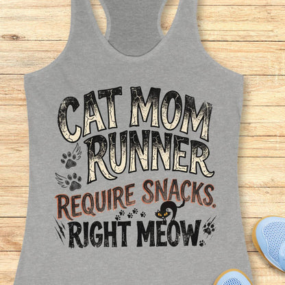 A heather gray racerback tank top laid flat on a light wood background, featuring a distressed graphic that reads 'CAT MOM RUNNER REQUIRE SNACKS. RIGHT MEOW' with paw prints, wings, a small cat silhouette, and partial blue shoes at the edges.
