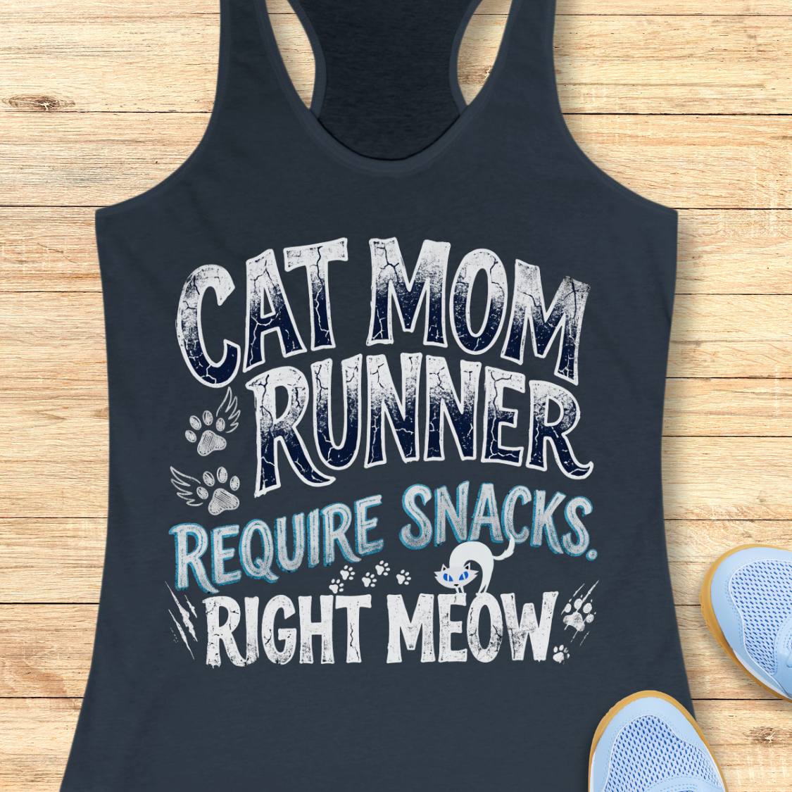 A indigo racerback tank top laid flat on a light wood background, featuring a humorous graphic that reads 'CAT MOM RUNNER REQUIRE SNACKS. RIGHT MEOW' in white and light blue text with paw prints and a small cat illustration.