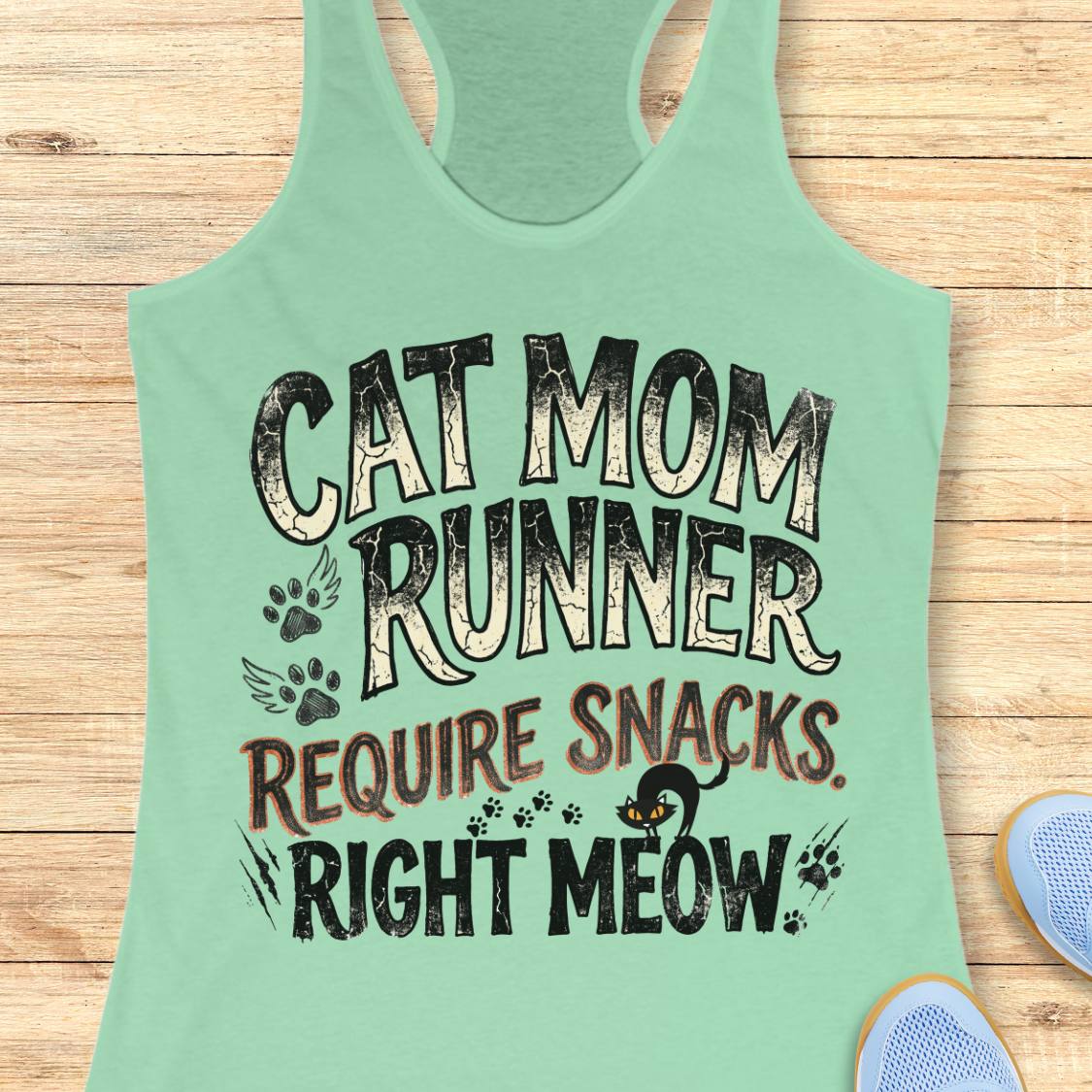 A mint green racerback tank top laid flat on a light wood background, featuring a distressed graphic that reads 'CAT MOM RUNNER REQUIRE SNACKS. RIGHT MEOW.