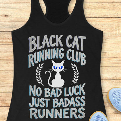 A black racerback tank top laid flat on a light wood background, featuring a graphic design with the text 'BLACK CAT RUNNING CLUB' and 'NO BAD LUCK JUST BADASS RUNNERS' around a white cat illustration with blue eyes.