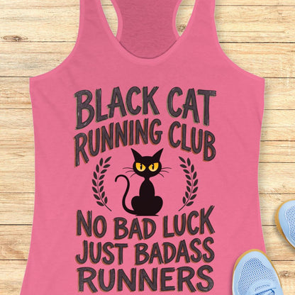 A pink racerback tank top laid flat on a light wood surface, featuring a black cat graphic with yellow eyes and the text 'BLACK CAT RUNNING CLUB' and 'NO BAD LUCK JUST BADASS RUNNERS.' Partial blue sneakers are visible at the edges as styling props.
