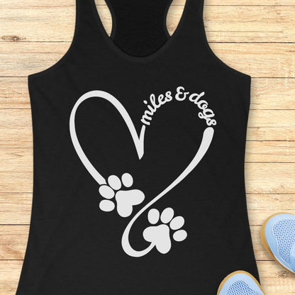 A black racerback tank top laid flat on a light wood surface, featuring a white graphic of a heart formed by a running path and paw prints, with the words 'miles & dogs' curved over the top. Part of a blue shoe is visible in the bottom right corner.
