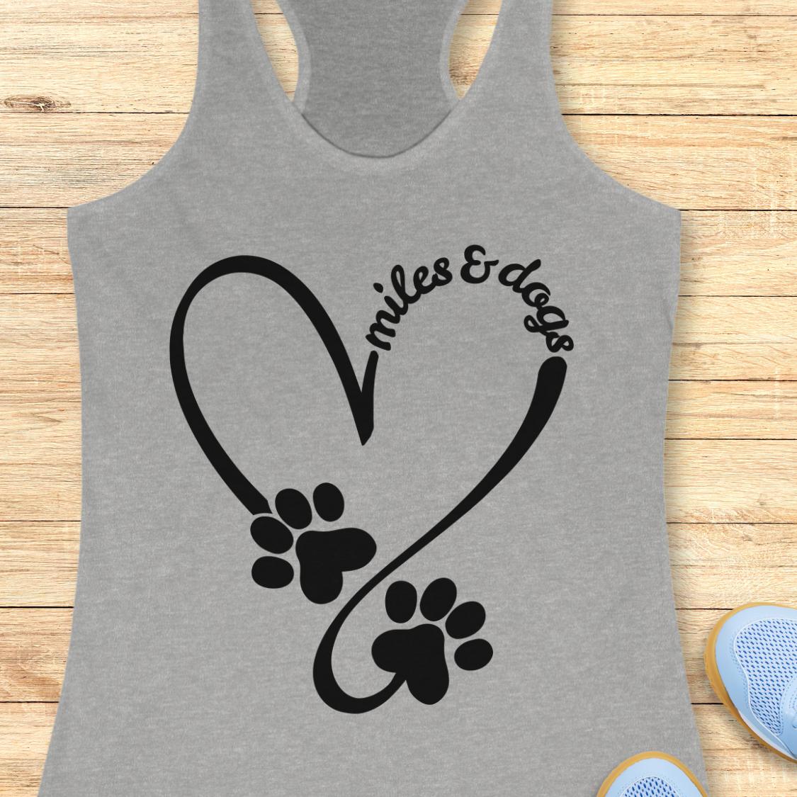 A heather gray racerback tank top laid flat on a light wood surface, featuring a black graphic of a heart shape formed by a loop and swoosh with two paw prints and the words 'miles & dogs' in script.