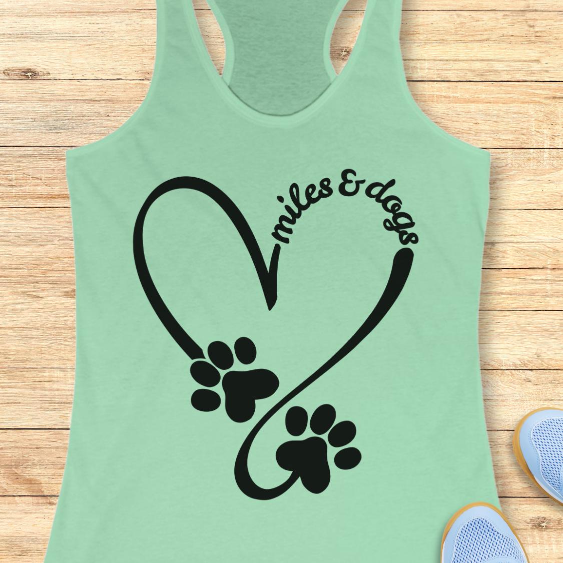 A mint green racerback tank top laid flat on a light wood background, featuring a black heart outline with two paw prints and the words 'miles & dogs' curved above them.