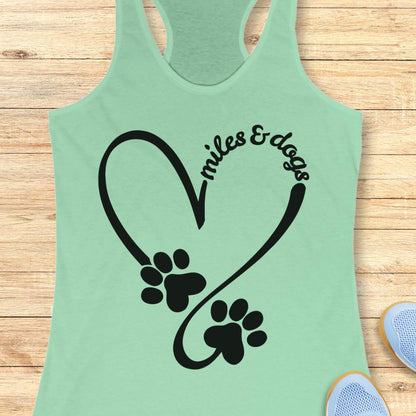 A mint green racerback tank top laid flat on a light wood background, featuring a black heart outline with two paw prints and the words 'miles & dogs' curved above them.