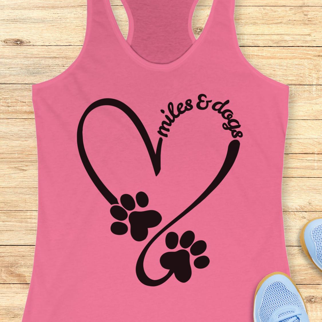 A pink racerback tank top laid flat on a light wood surface, featuring a black heart outline with two dog paw prints and the words 'miles & dogs' curved over one of the paws. Partial blue sneakers are visible at the bottom right as styling props.