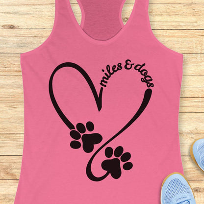A pink racerback tank top laid flat on a light wood surface, featuring a black heart outline with two dog paw prints and the words 'miles & dogs' curved over one of the paws. Partial blue sneakers are visible at the bottom right as styling props.