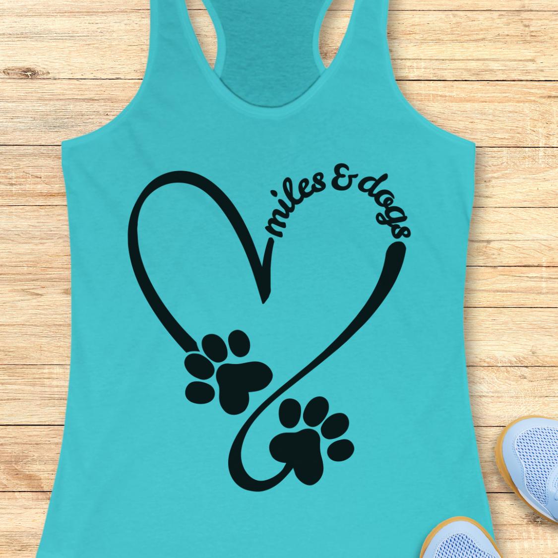 A turquoise racerback tank top laid flat on a light wood surface, featuring a black heart outline with two paw prints and the words 'miles & dogs' curved above them. Partial shoes are visible at the bottom right as styling props.