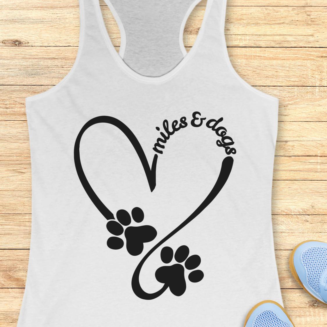 A white racerback tank top laid flat on a light wood surface, featuring a black heart outline with two paw prints and the words 'miles & dogs' curved over the top. Partial blue shoes are visible at the bottom right as styling props.