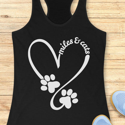 A black racerback tank top laid flat on a light wood surface, featuring a white heart-shaped graphic made from paw prints and the words 'miles & cats.' Partial blue shoes are visible at the bottom right as styling props.