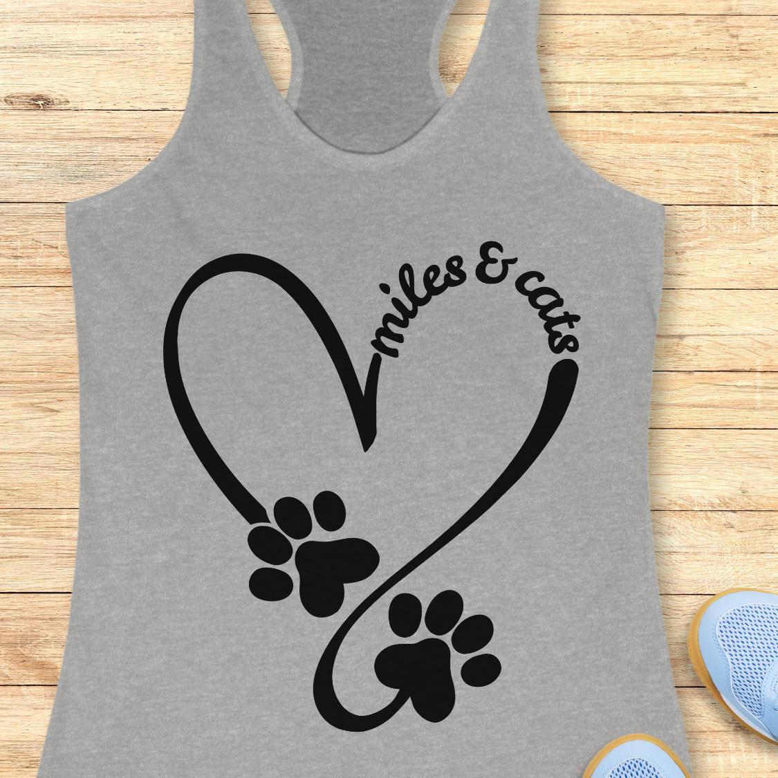 A heather gray racerback tank top laid flat on a light wood background, featuring a black graphic of a heart made from paw prints and the words 'miles & cats' in script.