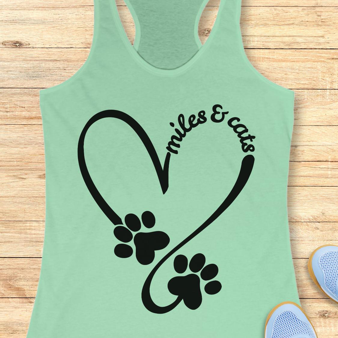 A mint green racerback tank top laid flat on a light wood background, featuring a black graphic of a heart outline with two paw prints and the words 'smiles & cats'.