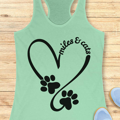 A mint green racerback tank top laid flat on a light wood background, featuring a black graphic of a heart outline with two paw prints and the words 'smiles & cats'.