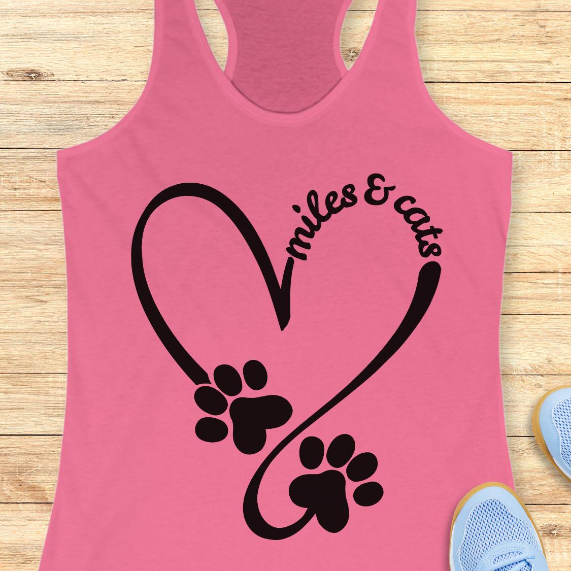 A pink racerback tank top with a black graphic of a heart, paw prints, and the text 'smiles & cats', displayed flat on a light wood background with partial blue sneakers visible at the edges.