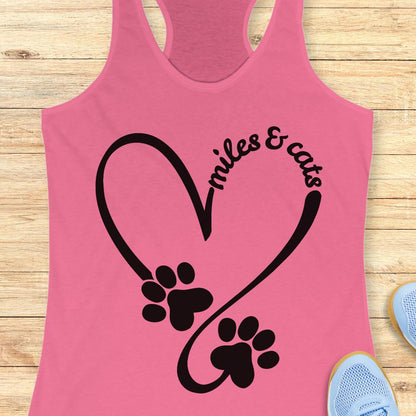 A pink racerback tank top with a black graphic of a heart, paw prints, and the text 'smiles & cats', displayed flat on a light wood background with partial blue sneakers visible at the edges.