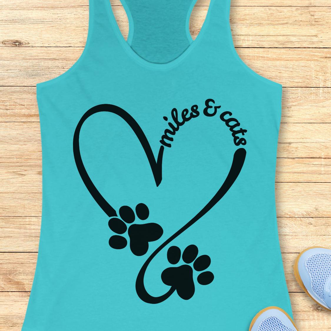 A turquoise racerback tank top laid flat on a light wood surface, featuring a black heart outline with two paw prints and the words 'smiles & cats' in script. Part of a blue shoe is visible at the bottom right.