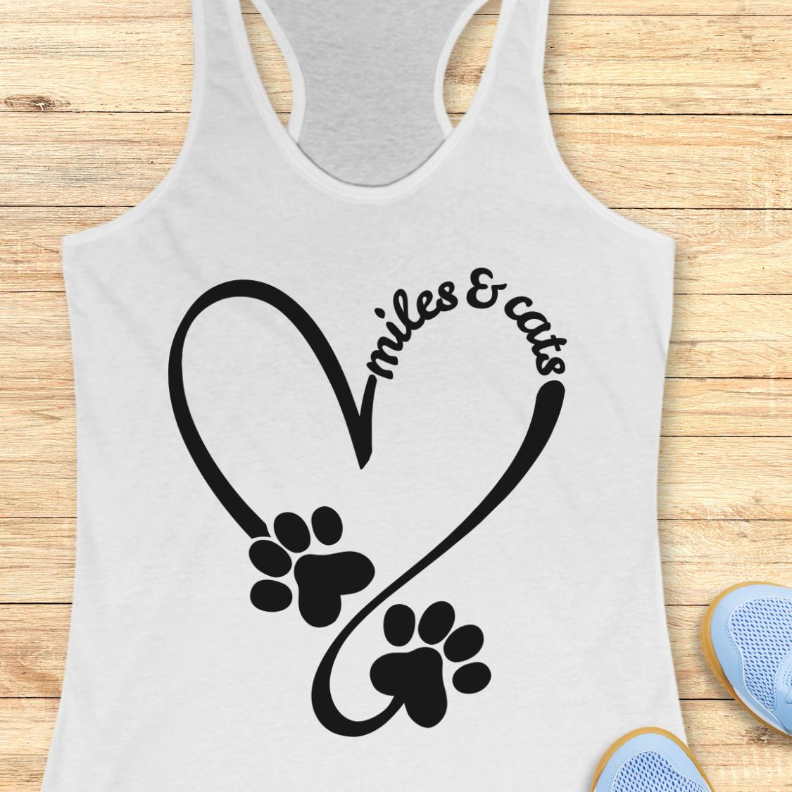 A white racerback tank top laid flat on a light wood surface, featuring a black graphic of a heart made from a running path and two paw prints, with the words 'miles & cats' curved along the top right of the heart. Partial blue shoes are visible at the bottom right edge.