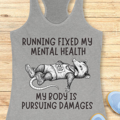 A gray racerback tank top laid flat on a light wood surface, featuring a black line-art illustration of a running opossum wearing a race bib and the text 'RUNNING FIXED MY MENTAL HEALTH' and 'MY BODY IS PURSUEING DAMAGES'.
