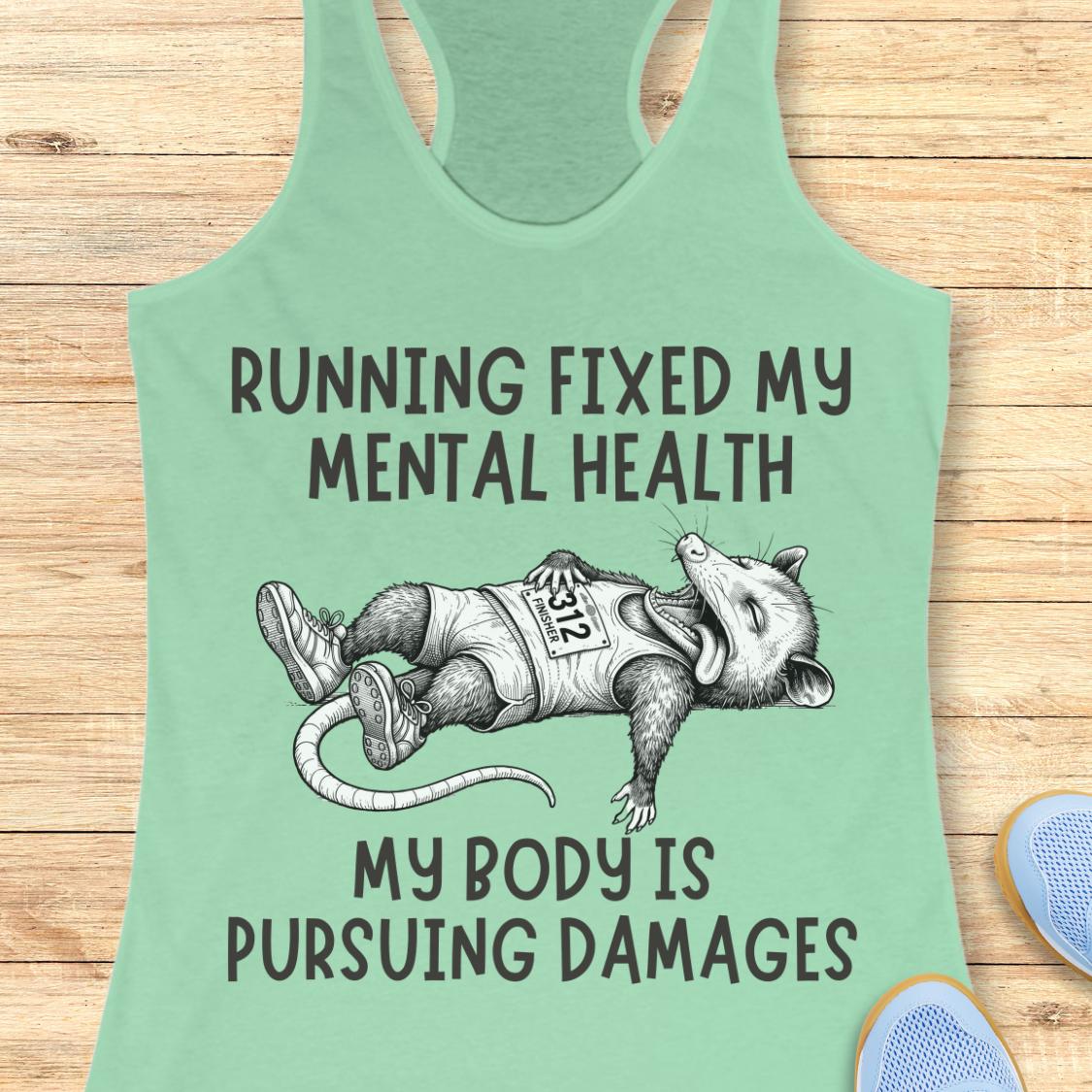 A mint green racerback tank top laid flat on a light wood background, featuring a humorous graphic of a raccoon wearing a 'RSP' vest and the text 'RUNNING FIXED MY MENTAL HEALTH MY BODY IS PURSUEING DAMAGES'.