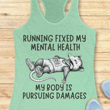 A mint green racerback tank top laid flat on a light wood background, featuring a humorous graphic of a raccoon wearing a 'RSP' vest and the text 'RUNNING FIXED MY MENTAL HEALTH MY BODY IS PURSUEING DAMAGES'.