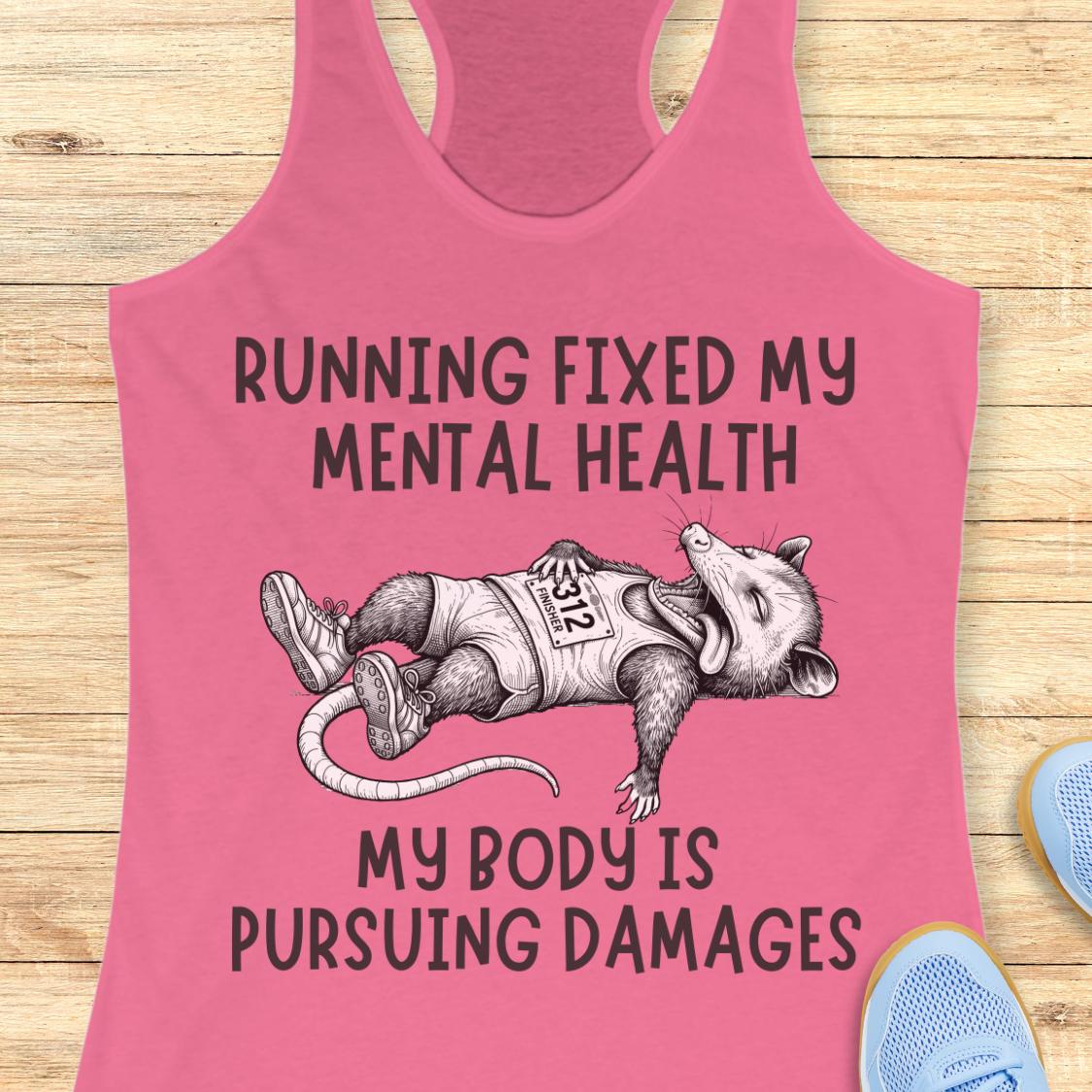 A pink racerback tank top laid flat on a light wood background, featuring a black graphic of a sleeping raccoon wearing a race bib and the text 'RUNNING FIXED MY MENTAL HEALTH MY BODY IS PURSUEING DAMAGES'. Partial blue shoes are visible at the bottom right edge.
