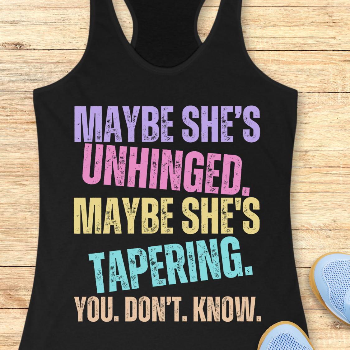 A black racerback tank top laid flat on a light wood surface, featuring distressed multicolor text that reads, 'MAYBE SHE'S UNHINGED. MAYBE SHE'S TAPERING.