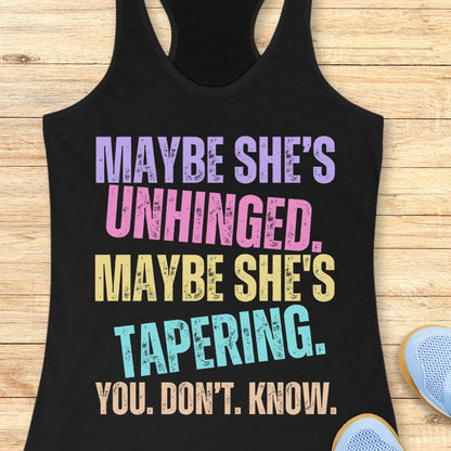 A black racerback tank top laid flat on a light wood surface, featuring distressed multicolor text that reads, 'MAYBE SHE'S UNHINGED. MAYBE SHE'S TAPERING.