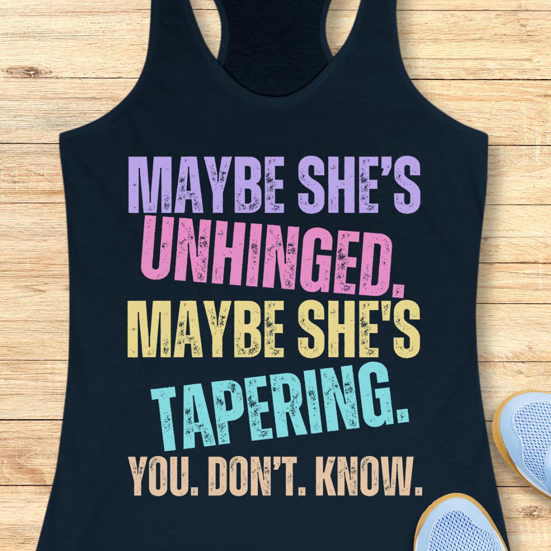 A navy racerback tank top laid flat on a light wood background, featuring a colorful distressed text graphic that reads, 'MAYBE SHE'S UNHINGED. MAYBE SHE'S TAPERING.