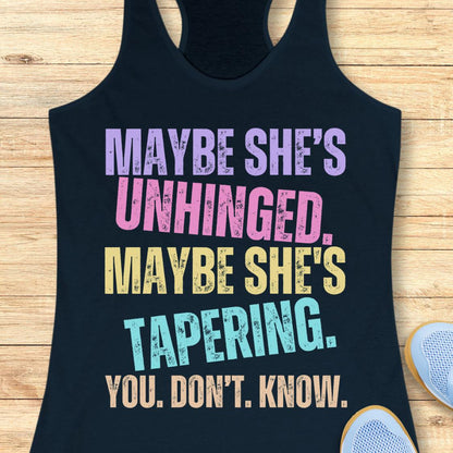 A navy racerback tank top laid flat on a light wood background, featuring a colorful distressed text graphic that reads, 'MAYBE SHE'S UNHINGED. MAYBE SHE'S TAPERING.