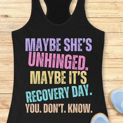 A black racerback tank top with a colorful distressed text graphic reading 'MAYBE SHE'S UNHINGED. MAYBE IT'S RECOVERY DAY.