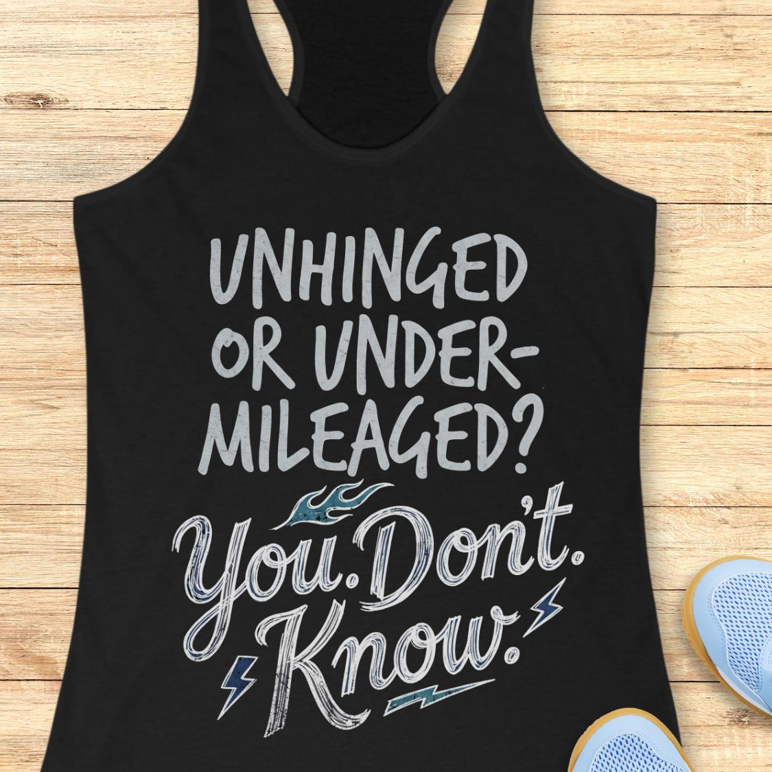 A black racerback tank top with a distressed white graphic text reading 'UNHINGED OR UNDER-MILEAGED? You Don't Know!' with small blue flame and lightning accents, laid flat on a light wood background with partial blue sneakers visible at the edges.