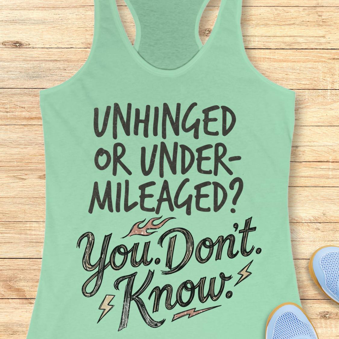 A mint green racerback tank top with a motivational graphic reading 'UNHINGED OR UNDER-MILEAGED? You Don't Know!' in dark lettering, laid flat on a light wood plank background with partial blue sneakers visible at the edges.