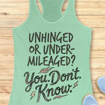 A mint green racerback tank top with a motivational graphic reading 'UNHINGED OR UNDER-MILEAGED? You Don't Know!' in dark lettering, laid flat on a light wood plank background with partial blue sneakers visible at the edges.