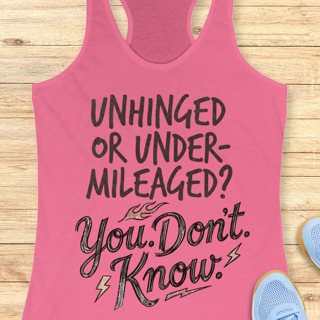 A pink racerback tank top laid flat on a light wood background, featuring the text 'UNHINGED OR UNDER-MILEAGED? You Don't Know.' in black lettering.