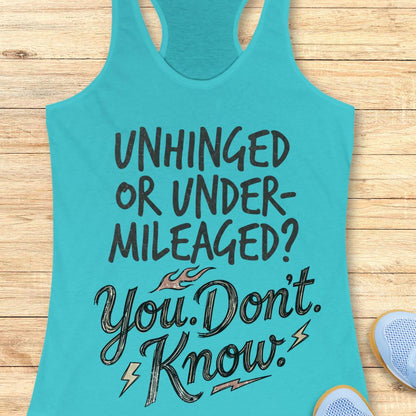 A turquoise racerback tank top with motivational text reading 'UNHINGED OR UNDER-MILEAGED? You Don't Know!' in black and gray lettering, laid flat on a light wood background with partial shoes visible at the edges.