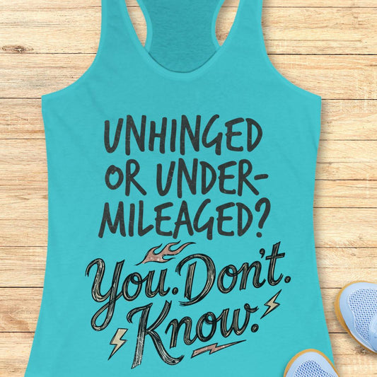 A turquoise racerback tank top with motivational text reading 'UNHINGED OR UNDER-MILEAGED? You Don't Know!' in black and gray lettering, laid flat on a light wood background with partial shoes visible at the edges.