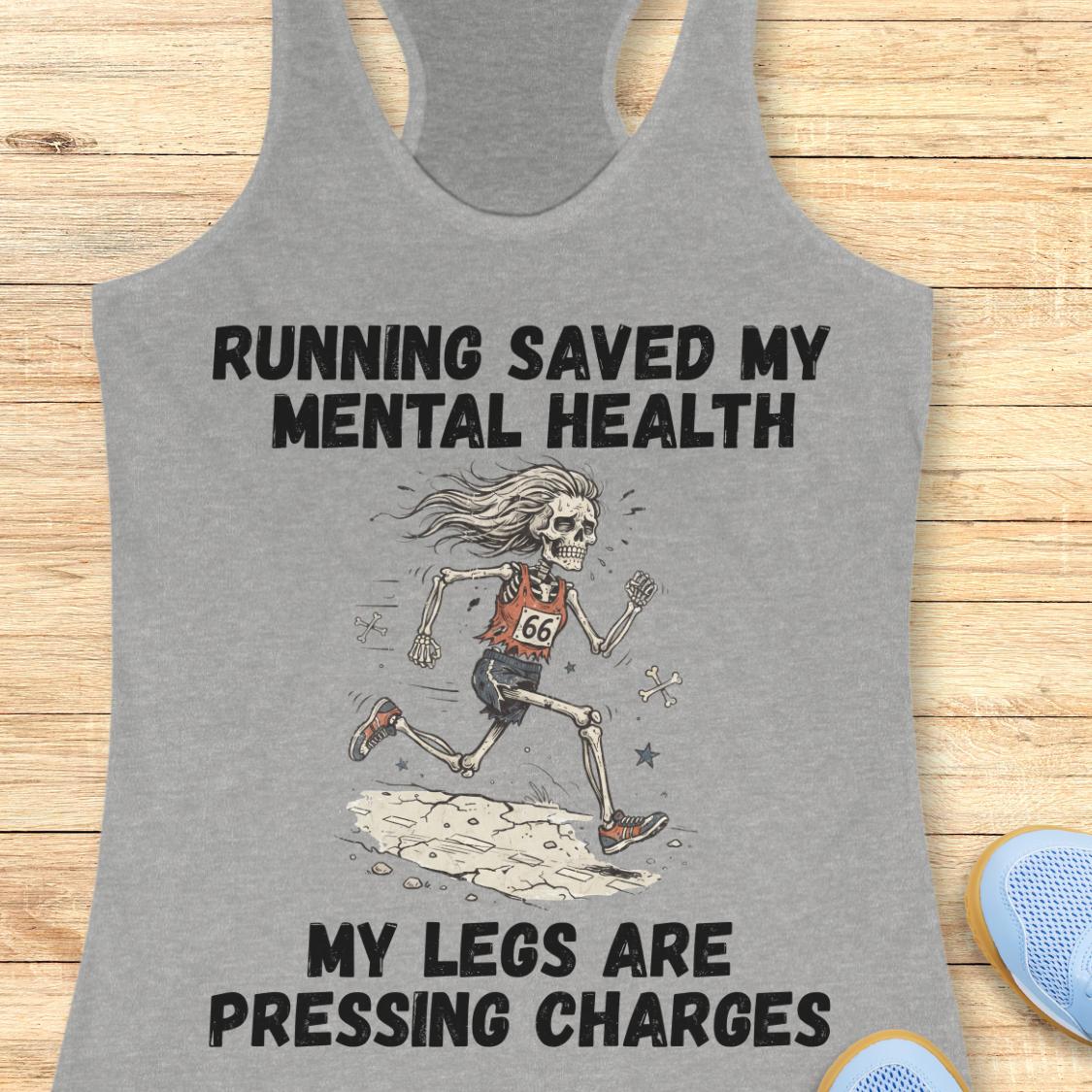 A heather gray racerback tank top laid flat on a light wood background, featuring a graphic of a running skeleton with the text 'RUNNING SAVED MY MENTAL HEALTH' above and 'MY LEGS ARE PRESSING CHARGES' below.