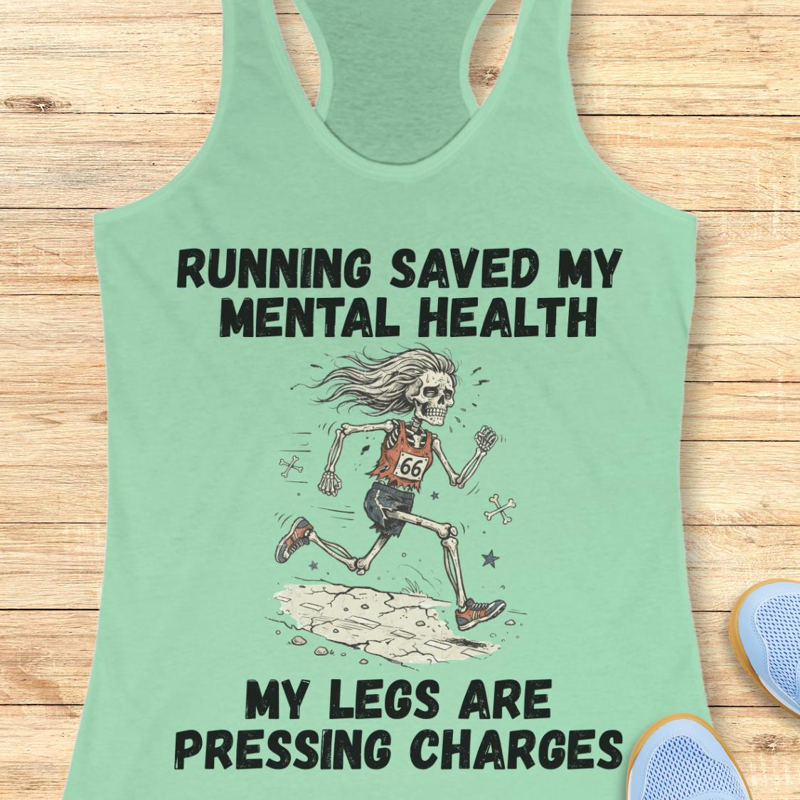 A mint green racerback tank top laid flat on a light wood background, featuring a humorous graphic of a running skeleton with the text 'RUNNING SAVED MY MENTAL HEALTH' and 'MY LEGS ARE PRESSING CHARGES'.