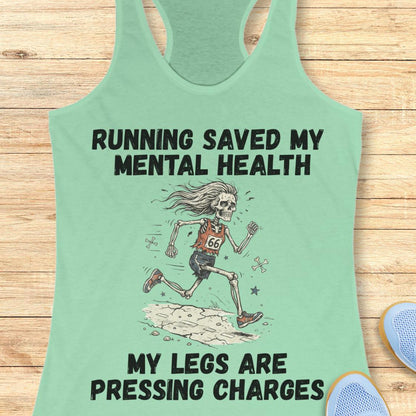 A mint green racerback tank top laid flat on a light wood background, featuring a humorous graphic of a running skeleton with the text 'RUNNING SAVED MY MENTAL HEALTH' and 'MY LEGS ARE PRESSING CHARGES'.
