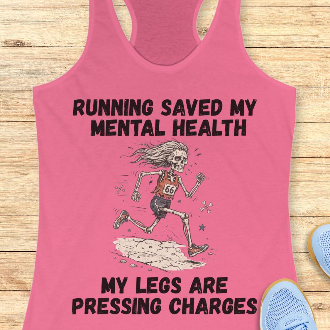 A pink racerback tank top laid flat on a light wood surface, featuring a black graphic of a running skeleton with the text 'RUNNING SAVED MY MENTAL HEALTH' above and 'MY LEGS ARE PRESSING CHARGES' below. Partial blue shoes are visible at the bottom right as styling props.