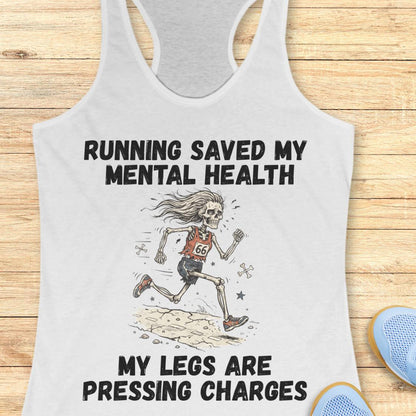 A white racerback tank top with a humorous graphic print featuring a running skeleton and the text 'RUNNING SAVED MY MENTAL HEALTH MY LEGS ARE PRESSING CHARGES', displayed flat on a light wood background with blue shoes partially visible in the corner.
