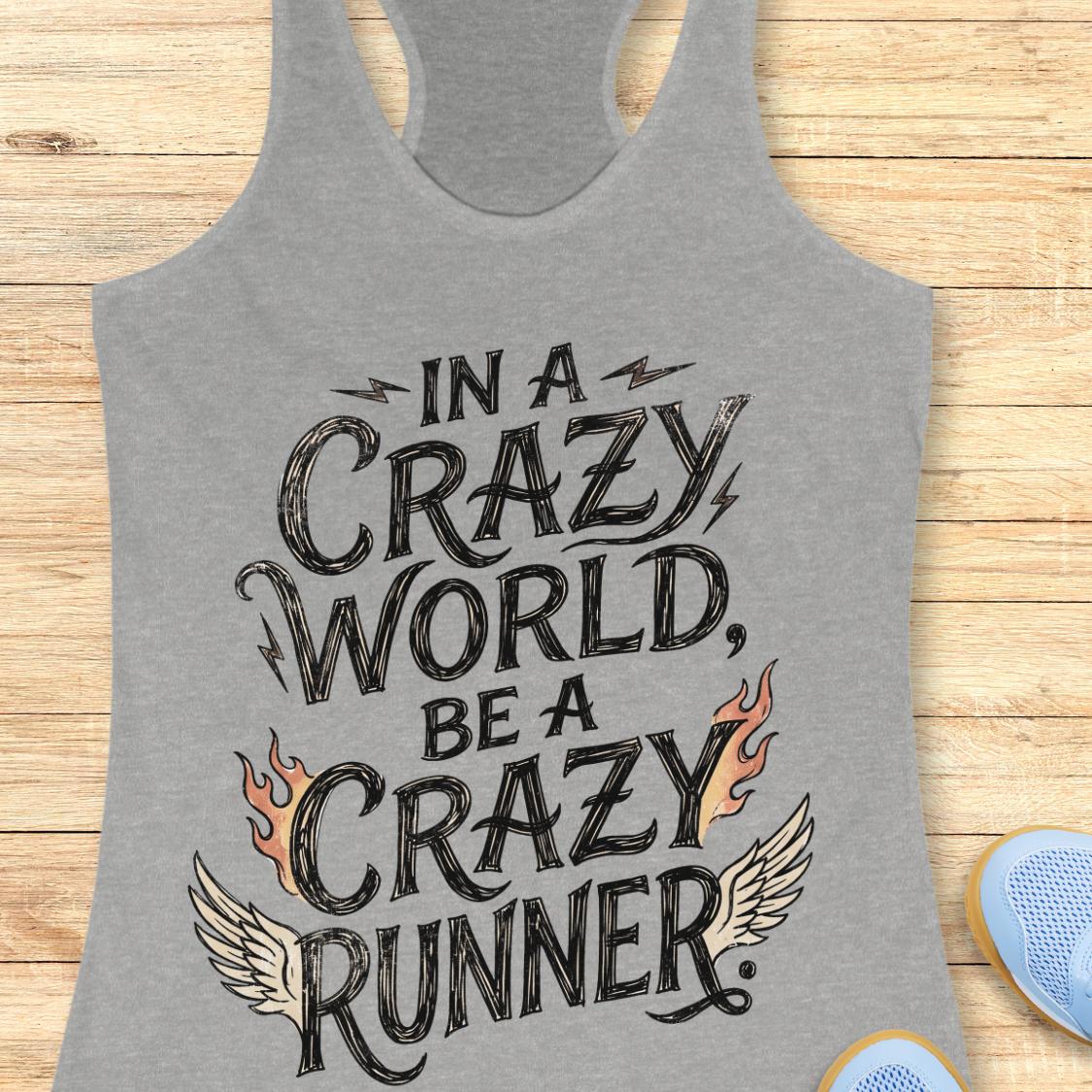 A heather gray racerback tank top with a graphic print reading 'IN A CRAZY WORLD, BE A CRAZY RUNNER' in black lettering, accented with flames and wings. The tank top is laid flat on a light wood background with partial blue sneakers visible at the bottom right.