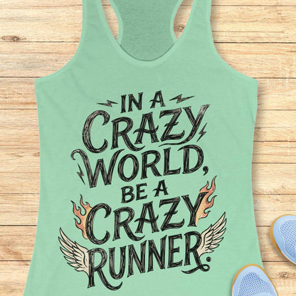 A mint green racerback tank top laid flat on a light wood plank background, featuring the graphic text 'IN A CRAZY WORLD, BE A CRAZY RUNNER' with black lettering, flame motifs, and wing details. Partial blue sneakers are visible at the bottom right as styling props.