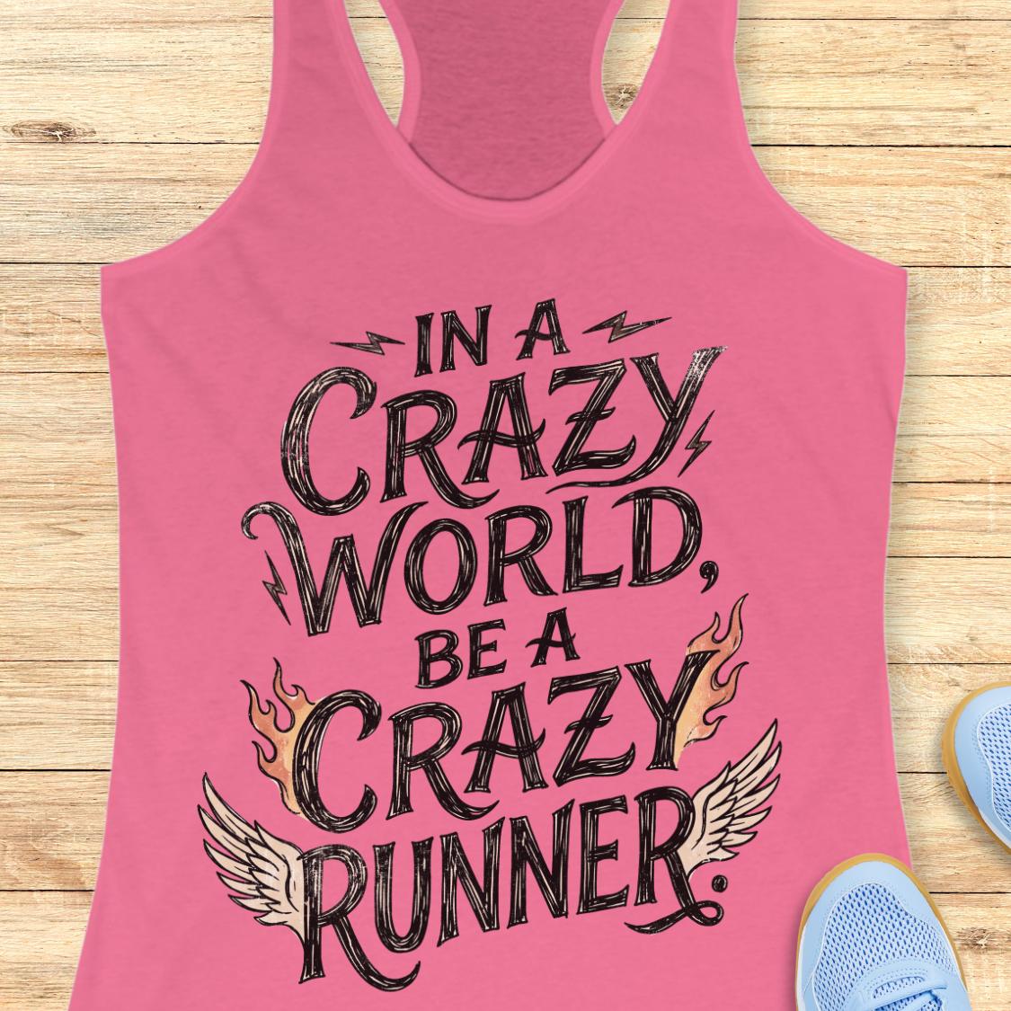 A pink racerback tank top laid flat on a light wood floor, featuring the phrase 'IN A CRAZY WORLD, BE A CRAZY RUNNER' in black lettering with flame and wing graphics. Partial blue sneakers are visible at the bottom right as styling props.