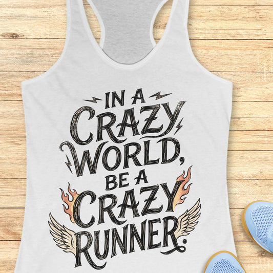 A white racerback tank top with a graphic print reading 'IN A CRAZY WORLD, BE A CRAZY RUNNER' in black lettering, accented with orange flames and beige wings, laid flat on a light wood background with partial blue shoes visible at the edges.