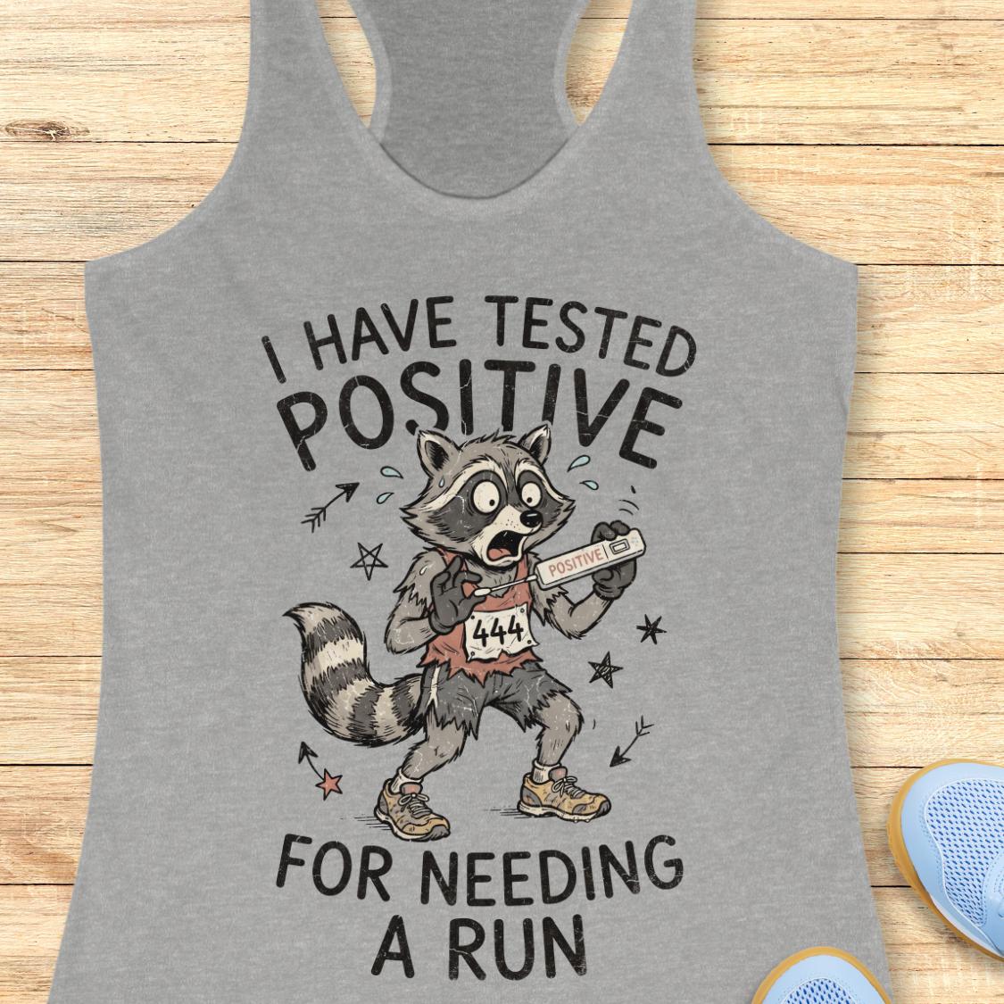 A heather gray racerback tank top laid flat on a light wood background, featuring a humorous graphic of a raccoon wearing a race bib and holding a finish line, with the text 'I HAVE TESTED POSITIVE FOR NEEDING A RUN'.